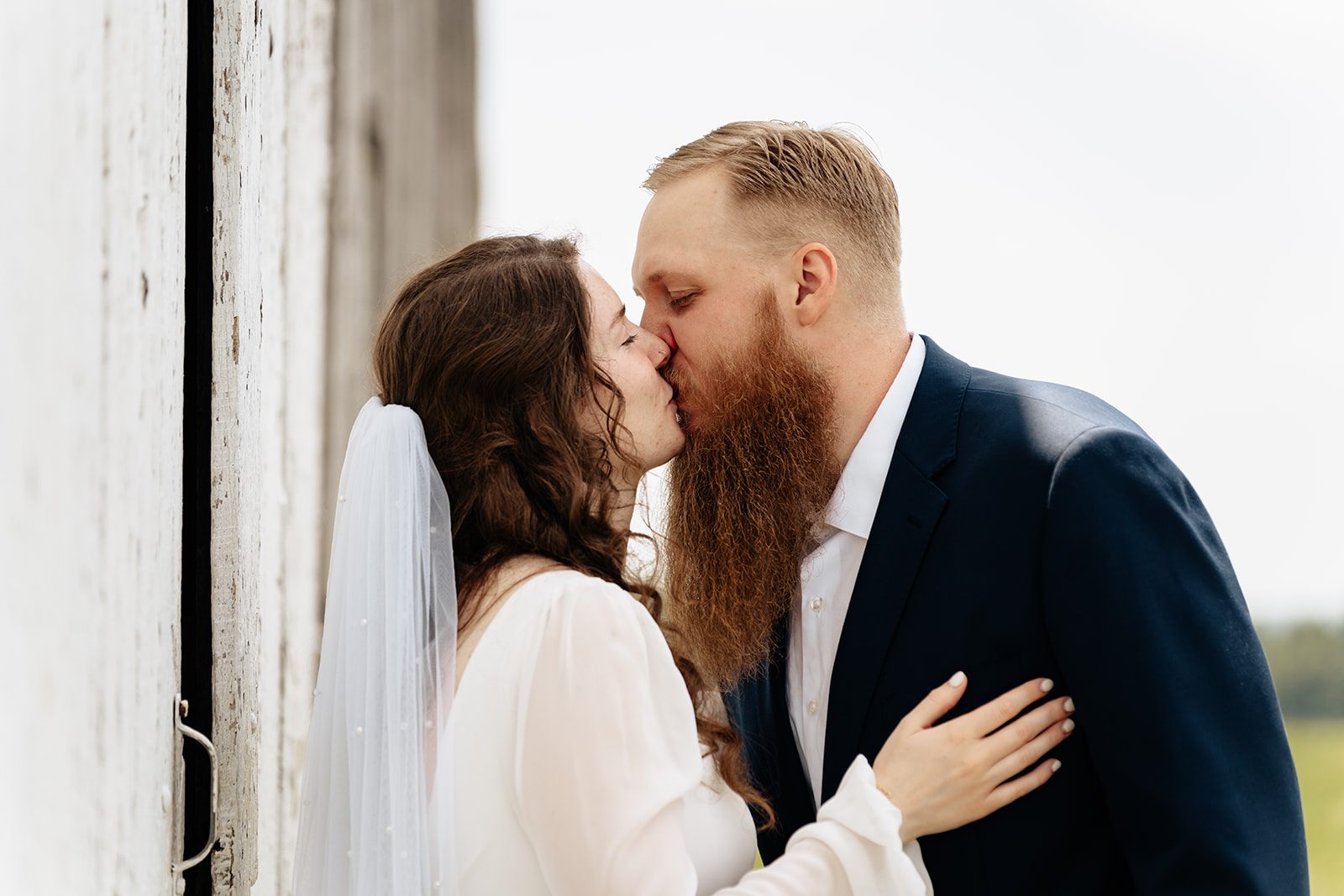 Bride and groom kissing in the open doorway of a weathered white barn with sunlit fields behind them