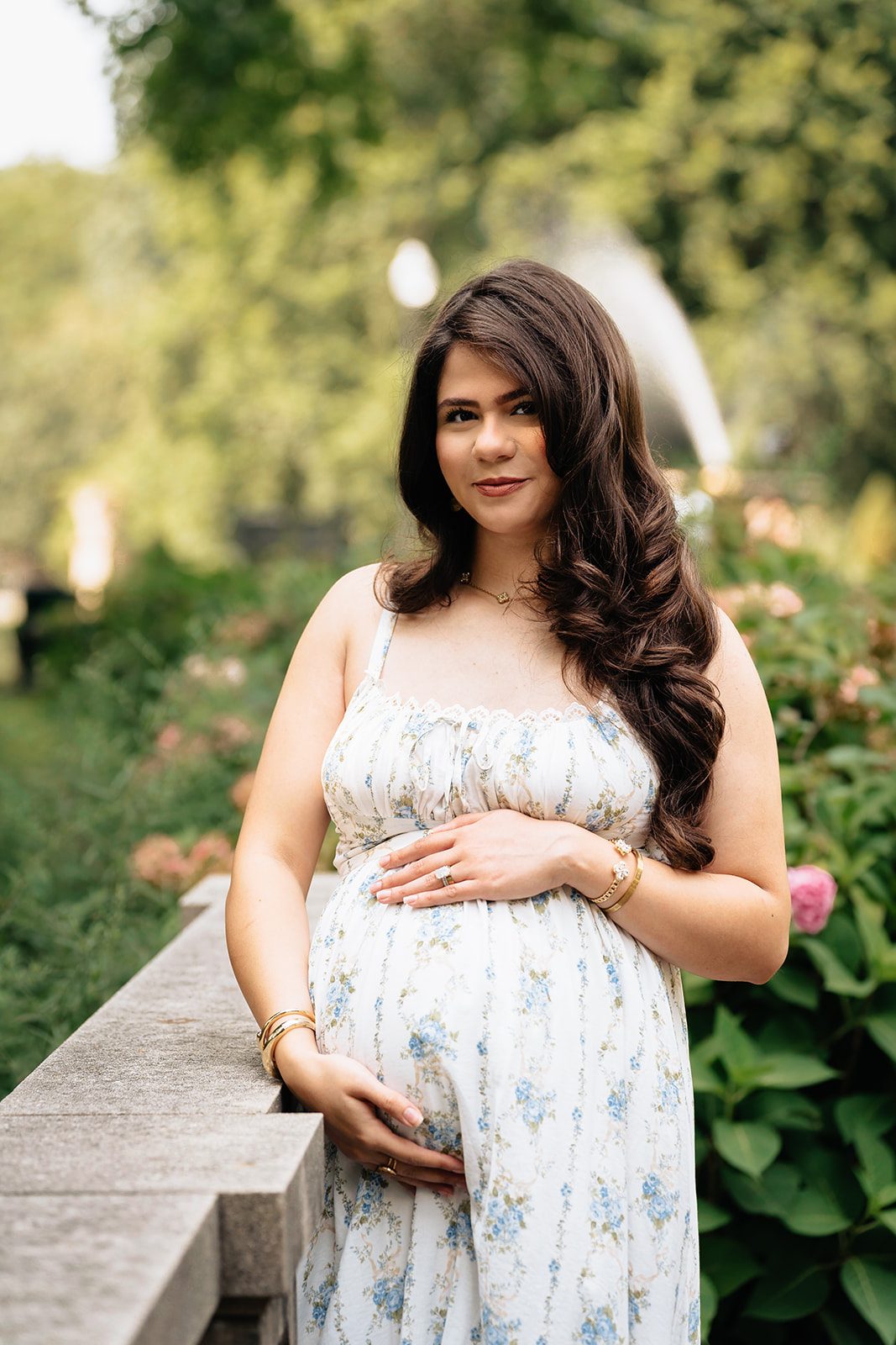 Hala in a blue floral dress resting a hand on her bump beside the carved stone balustrade of the Italian Cultural Gardens