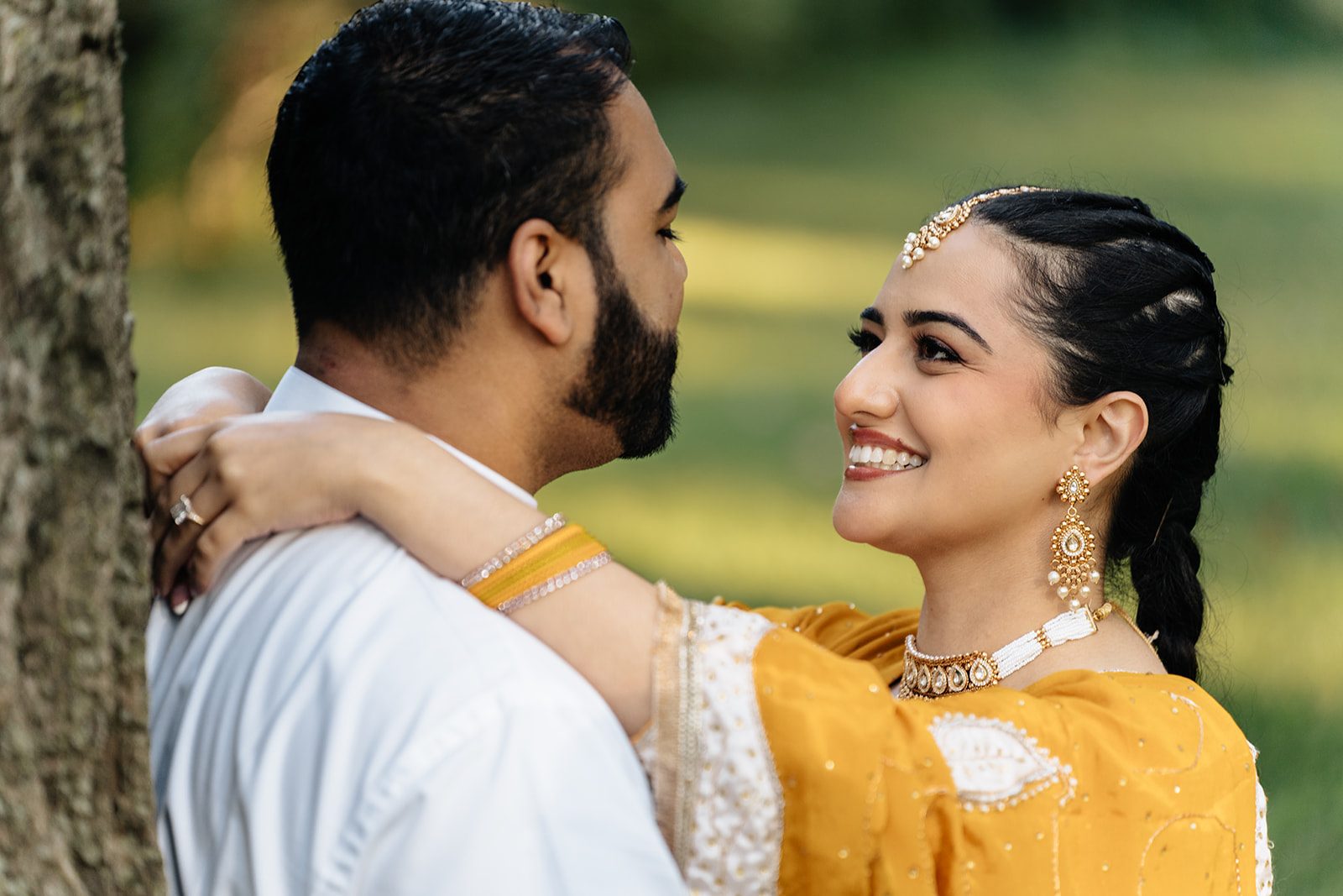 Close couple portrait with her arms around his neck, gold jewelry and maang tikka visible, both smiling genuinely