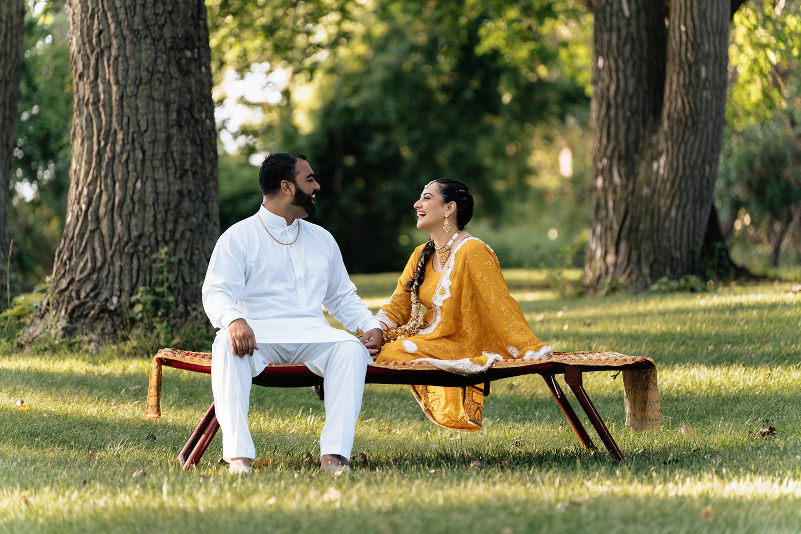 Couple seated at opposite ends of a woven charpai beneath huge mature tree trunks in dappled evening light