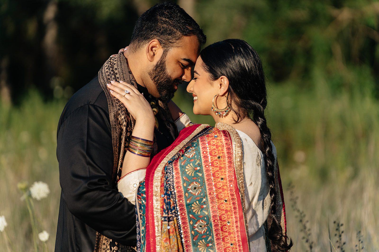 Couple resting their foreheads together in tall grass, striped dupatta and bangles catching the light among soft wildflowers