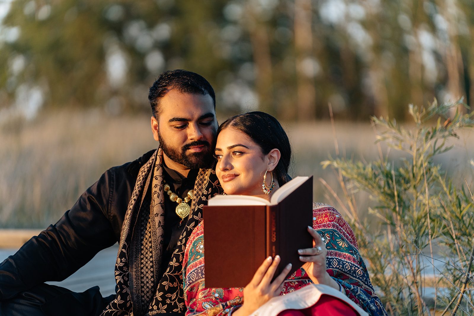 Ramneet and Harry reading a red leather-bound book together on a blanket, her head tucked into his shoulder
