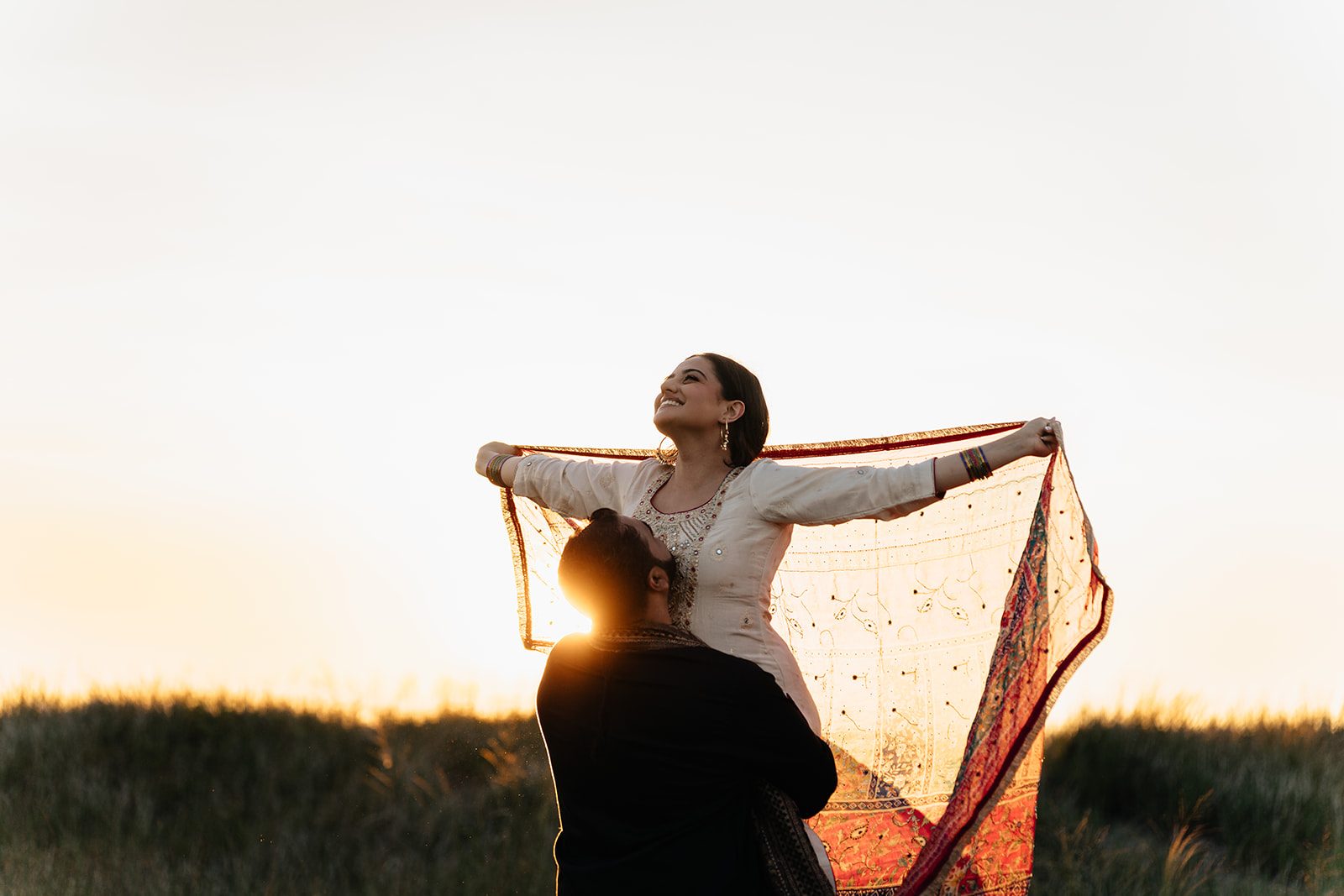 Ramneet standing in the dune grass with her dupatta opened wide as the sun shines through it, Harry nearly silhouetted below her