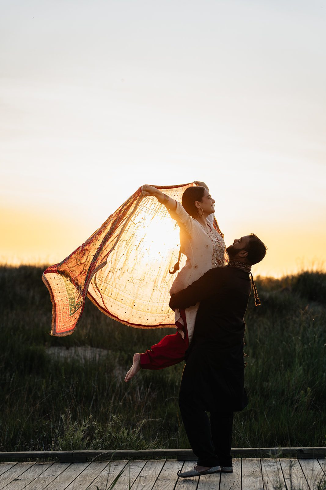 Harry lifting Ramneet on the boardwalk as her dupatta arcs overhead in the low sun