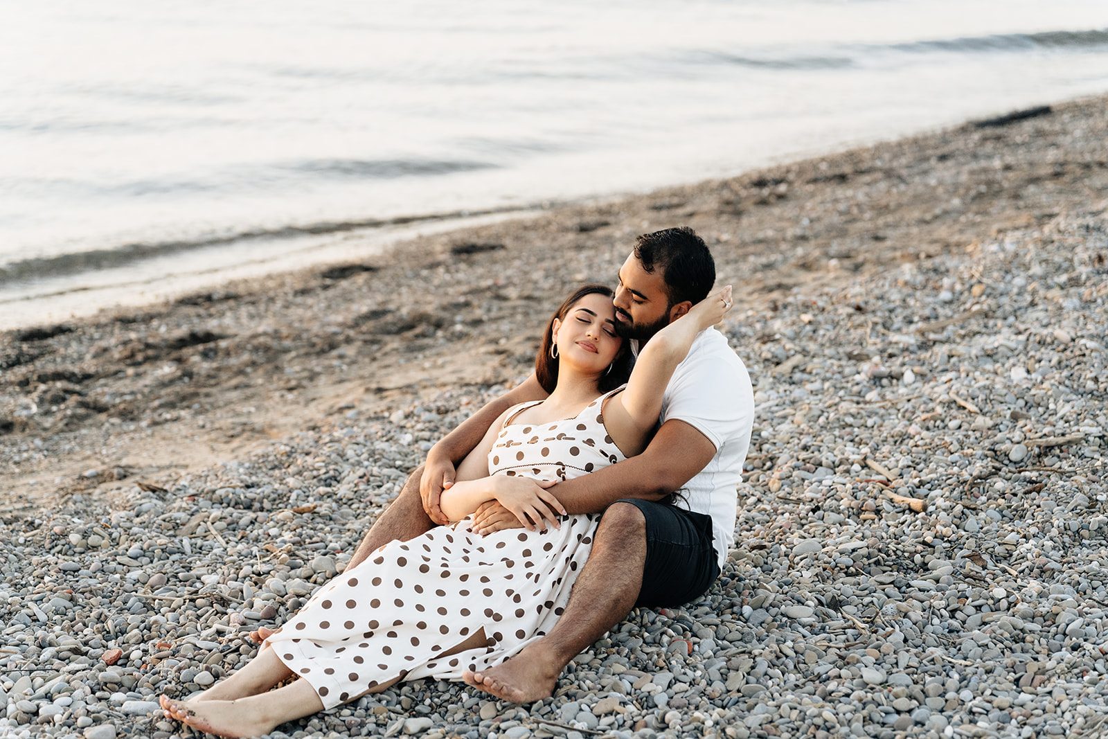 Couple sitting together on the pebbled shore, Ramneet leaning back into Harry with waves and the horizon behind them