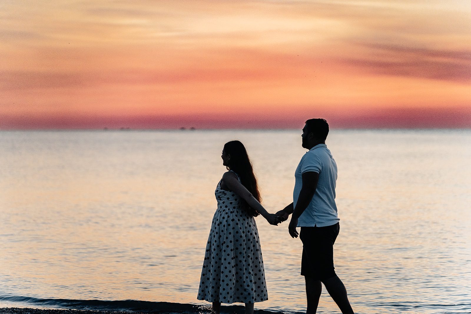 Couple small in the frame holding hands at the waterline beneath a wide pink and orange sunset sky