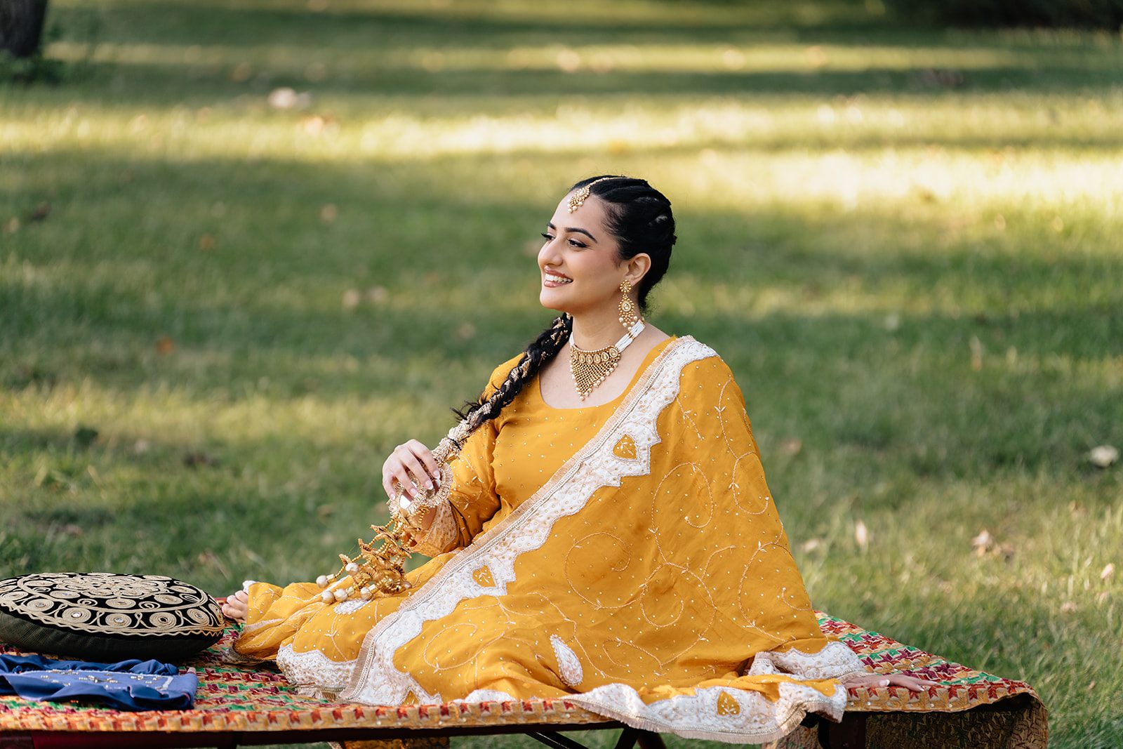 Ramneet seated alone on the woven charpai in a marigold-yellow suit, laughing softly