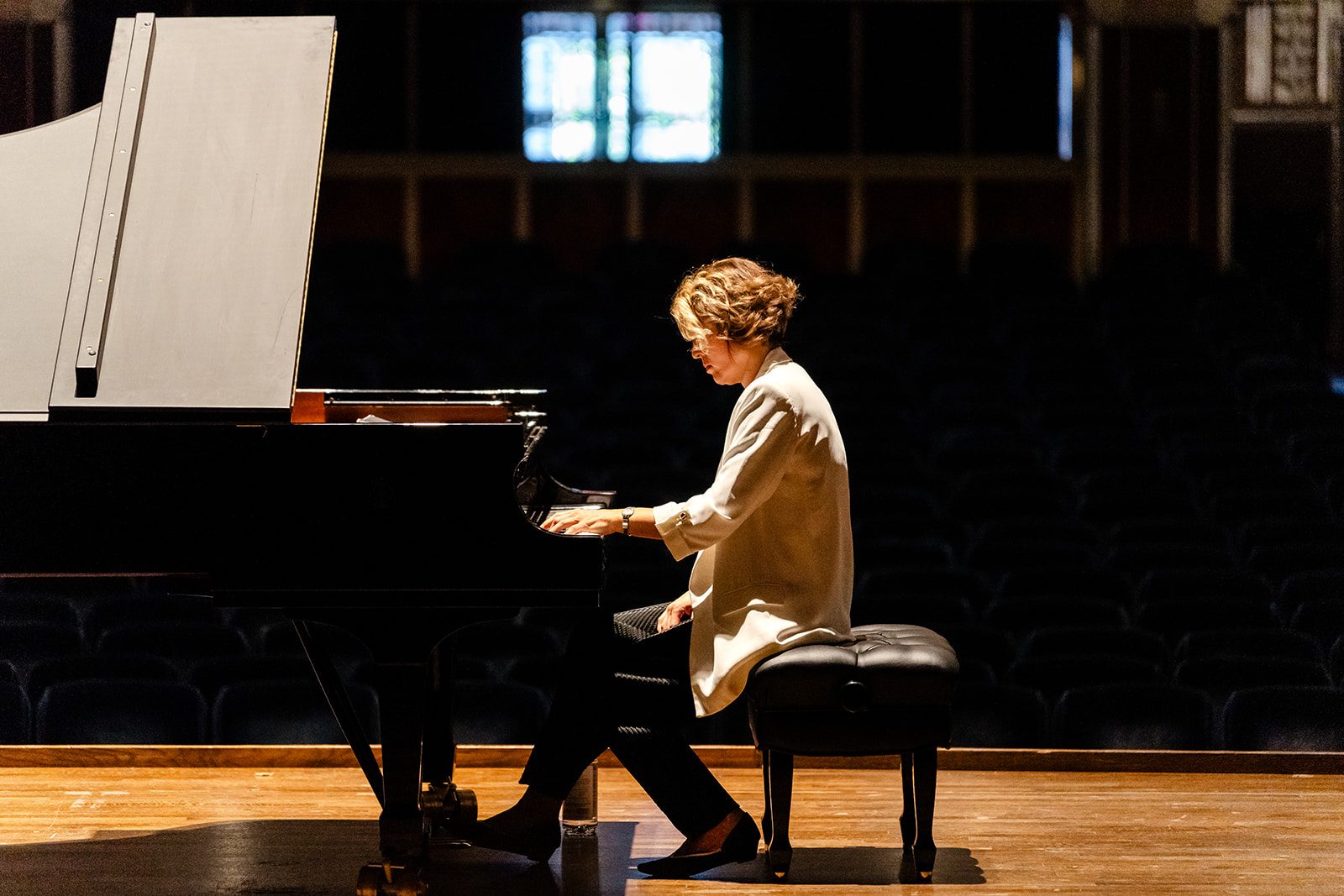 Clean side profile of the pianist at the keyboard in a white blazer with dark seats behind and a narrow stained-glass detail visible
