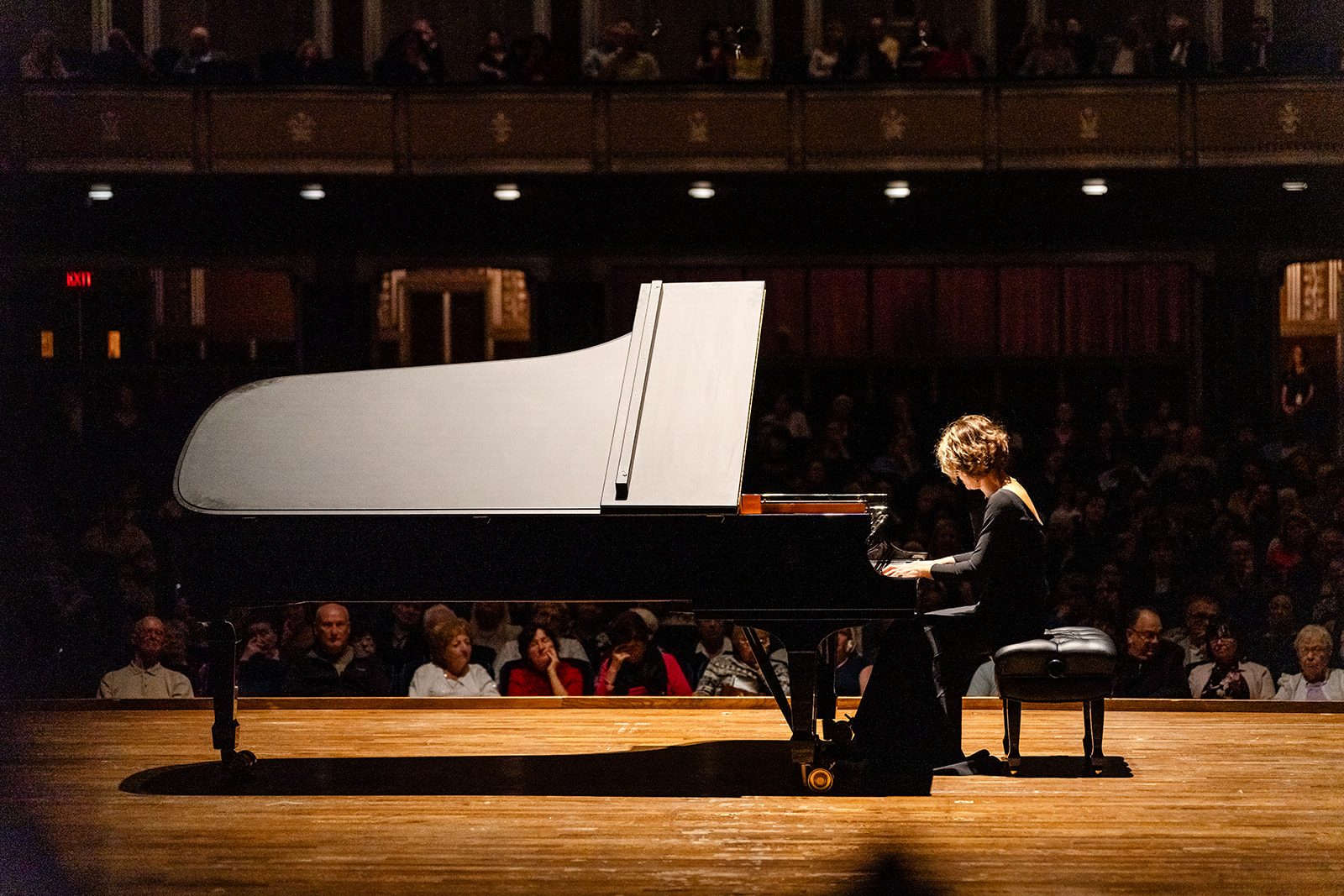 Side profile in a black gown at the piano with the full audience behind and the bright underside of the piano lid forming a graphic shape