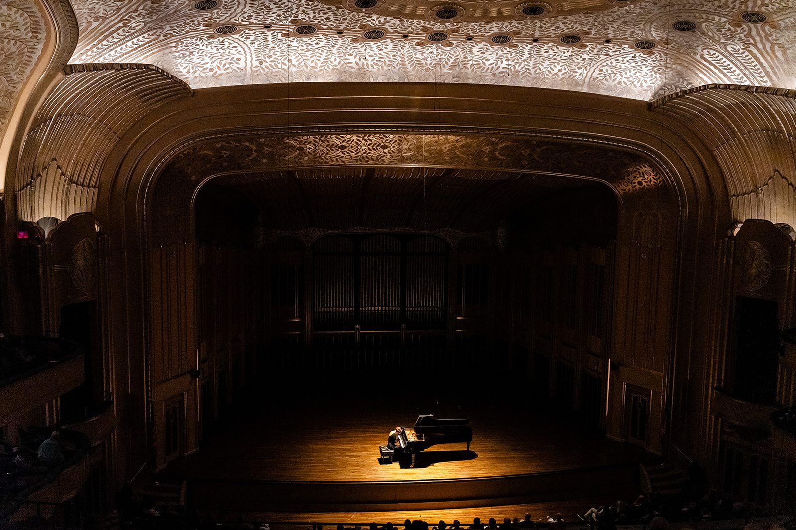 Balcony-wide view of Severance Music Center with the pianist and Steinway lit by a single warm pool of light beneath the ornate glowing ceiling