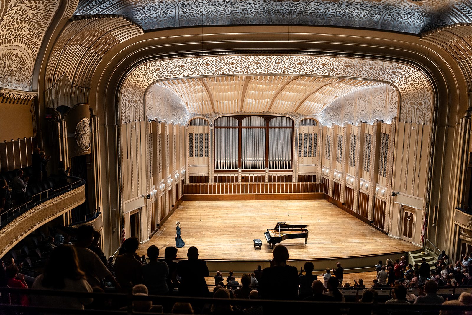 Full-house wide view at Severance Music Center as the performer enters before a seated audience, silhouetted heads in the foreground