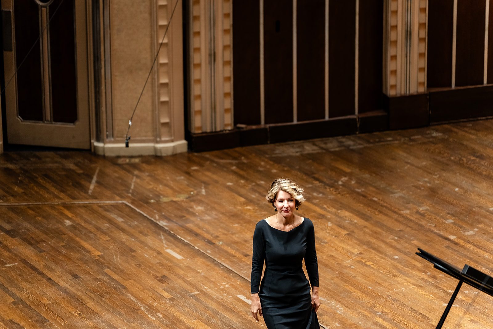 Overhead view of the pianist entering the stage in a black gown across a large expanse of warm wood floor