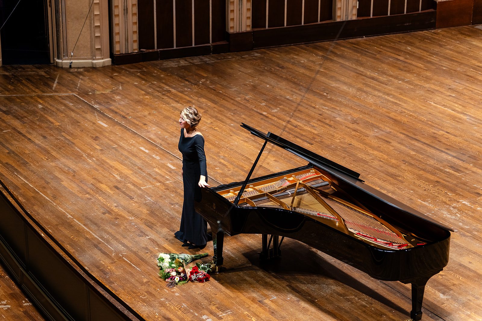 The pianist bowing beside the open piano with a hand to her chest and bouquets on the stage