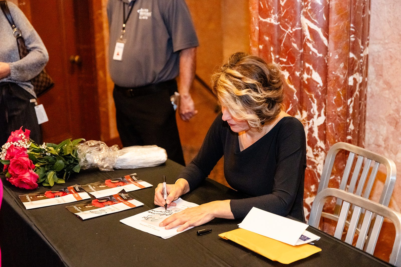 Warm lobby frame of the pianist signing a program at a table with roses nearby and red marble behind her